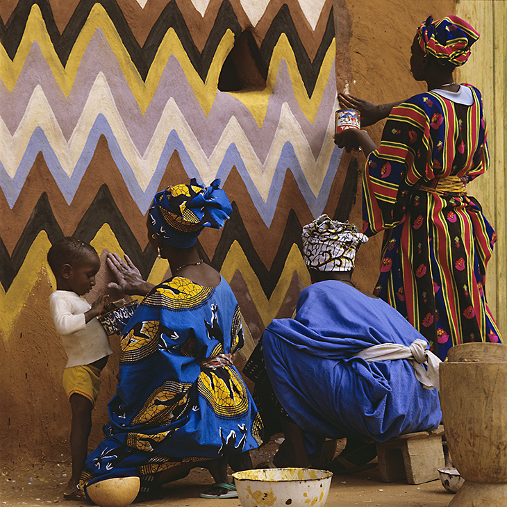 Soninke women divide a wall surface into panels before painting in Buanch, Mauritania, 1988. Photograph by Margaret Courtney-Clarke for African Canvas. Image shows two women kneeling and clad in blues, painting a wall. A third woman is standing and painting the wall. A child holds a paint pot. 